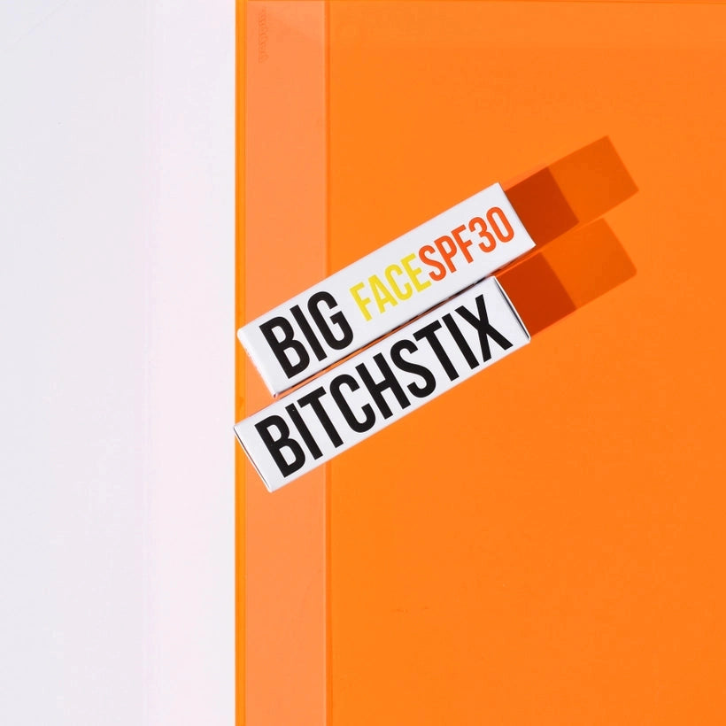 Two white product boxes, one labeled "BIG FACESPF30" featuring yellow and orange text and the other "BITCHSTIX" in bold black letters, are diagonally arranged on a vibrant orange and crisp white background with distinct shadows