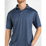 A man with a reddish beard models a dark blue performance polo shirt featuring a subtle repeating marlin pattern and white buttons, his hand casually in a pocket
