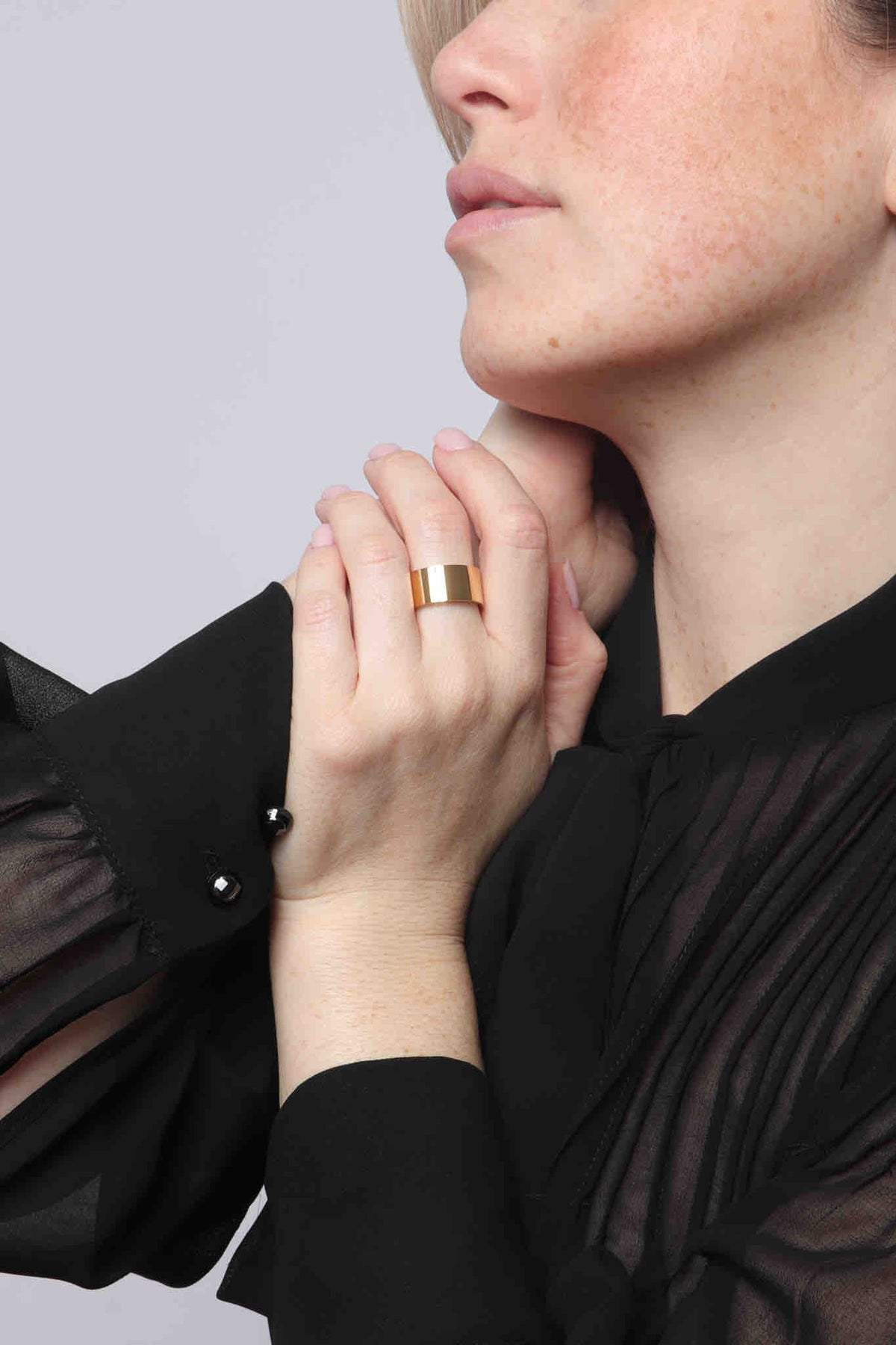 Close-up studio shot of a woman's hand with soft pink nail polish wearing a wide shiny gold band ring, her fingers delicately touching her neck, dressed in a black sheer pleated blouse with dark cuff buttons