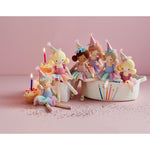 Six plush ballerina dolls with diverse skin tones and hair colors wearing pastel tiered tutus and sparkly party hats are arranged around cupcakes with lit candles and a woven basket embroidered with colorful candles on a glitter-scattered pink background