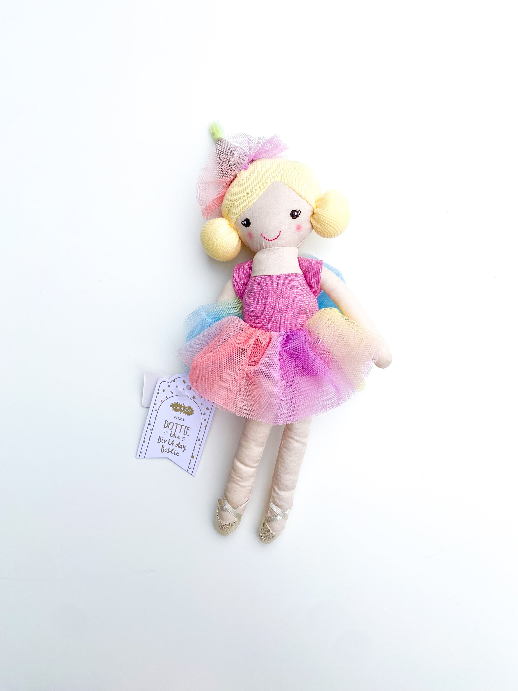 Mudpie plush toy Dottie the Birthday Bestie ballerina doll with blonde pigtails, a pink sparkly leotard, a vibrant rainbow tulle tutu, and gold ballet slippers, accompanied by its product tag