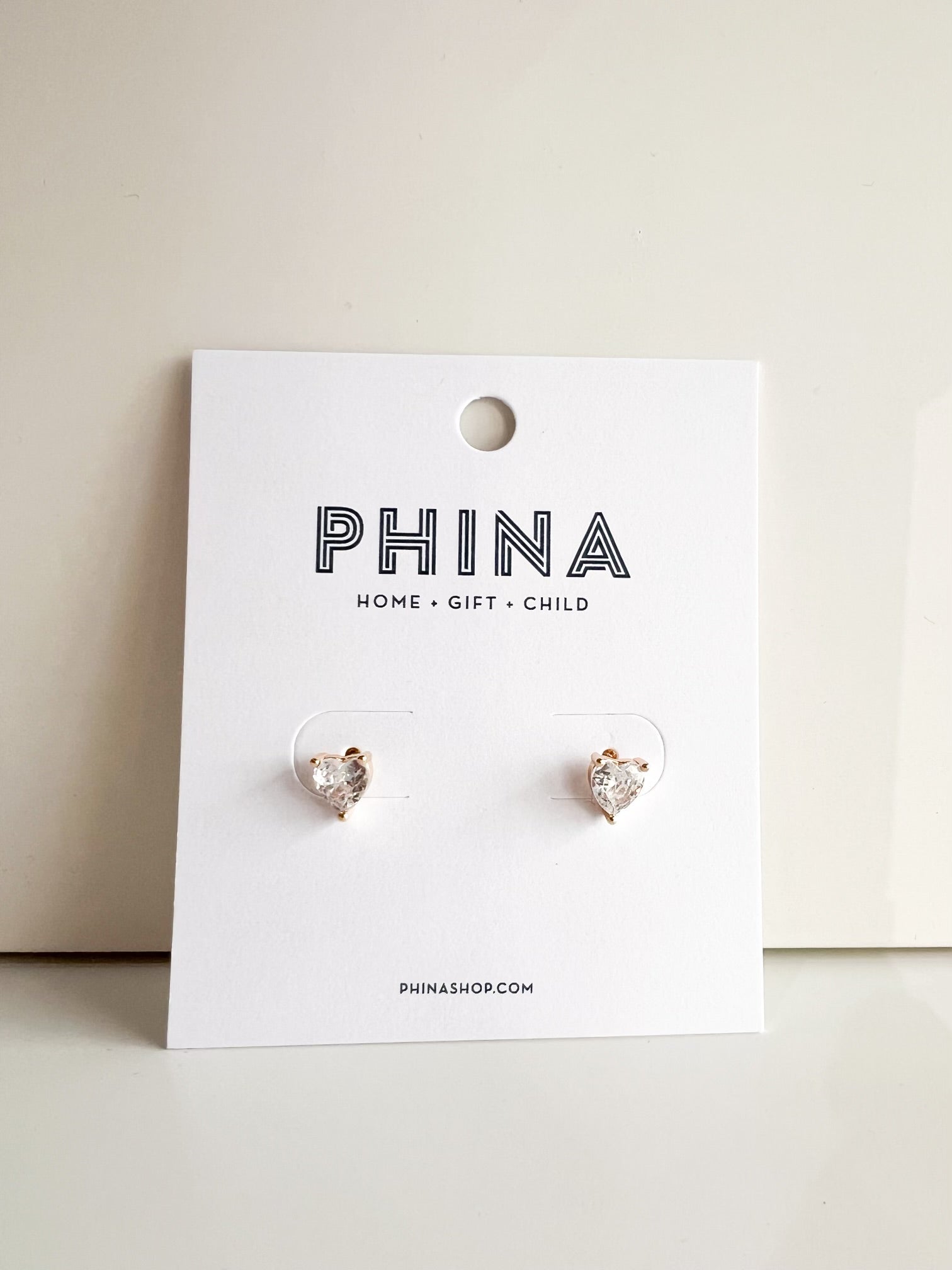 Gold heart shaped stud earrings with clear stones on a Phina branded display card