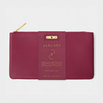 Product photo of a Katie Loxton burgundy faux leather January birthstone pouch with a gold zipper and a paper band detailing the Garnet stone and "Secret Message Inside"
