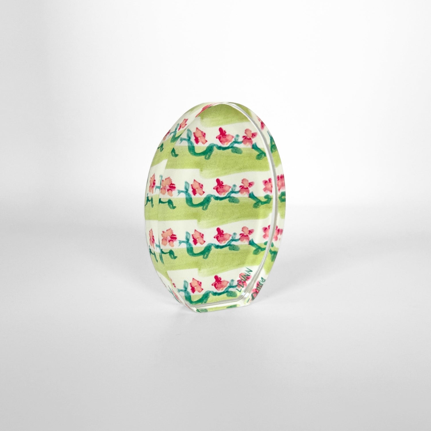 A glossy decorative oval object in a studio setting, adorned with wide green horizontal stripes and vibrant pink floral vine patterns with green leaves, bearing a faint LINN inscription