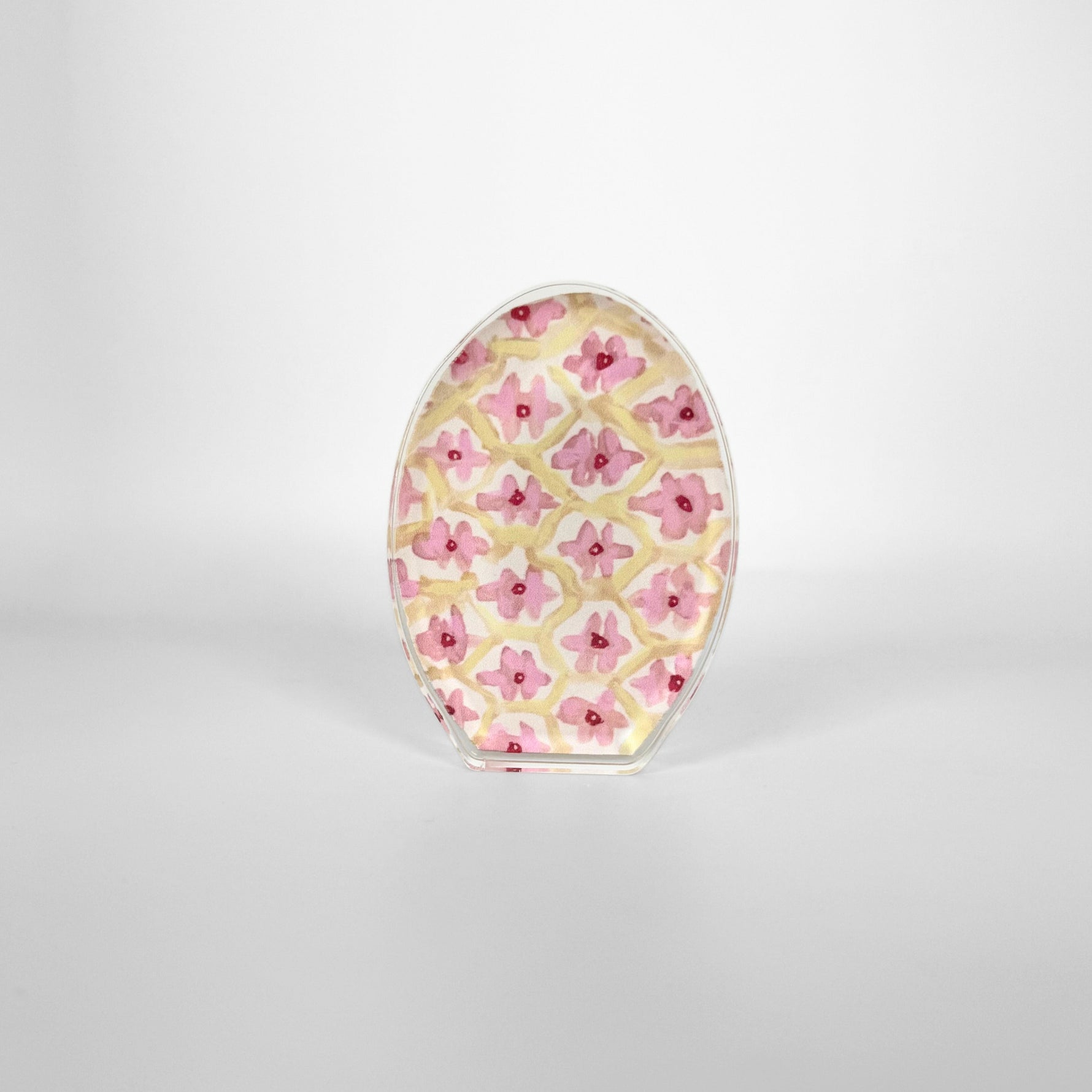 A clear oval decorative panel features a repeating pattern of light pink flowers with dark red centers interspersed within a pale yellow grid on a white studio background