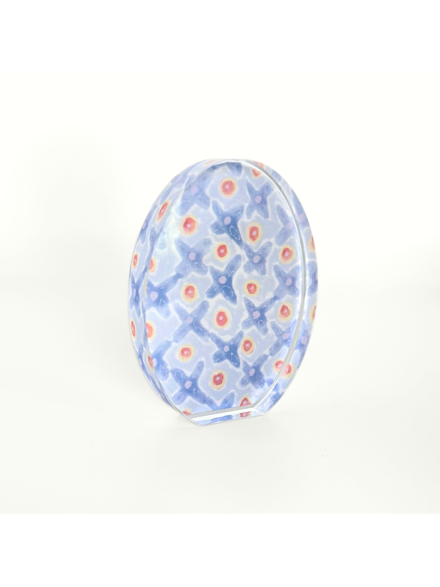 Clear glass dome paperweight featuring an internal pattern of light blue X-shapes interspersed with vibrant coral and yellow dots against a pale background