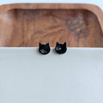 A pair of shiny black cat head stud earrings resting on a crisp white surface with a warm natural wooden bowl background