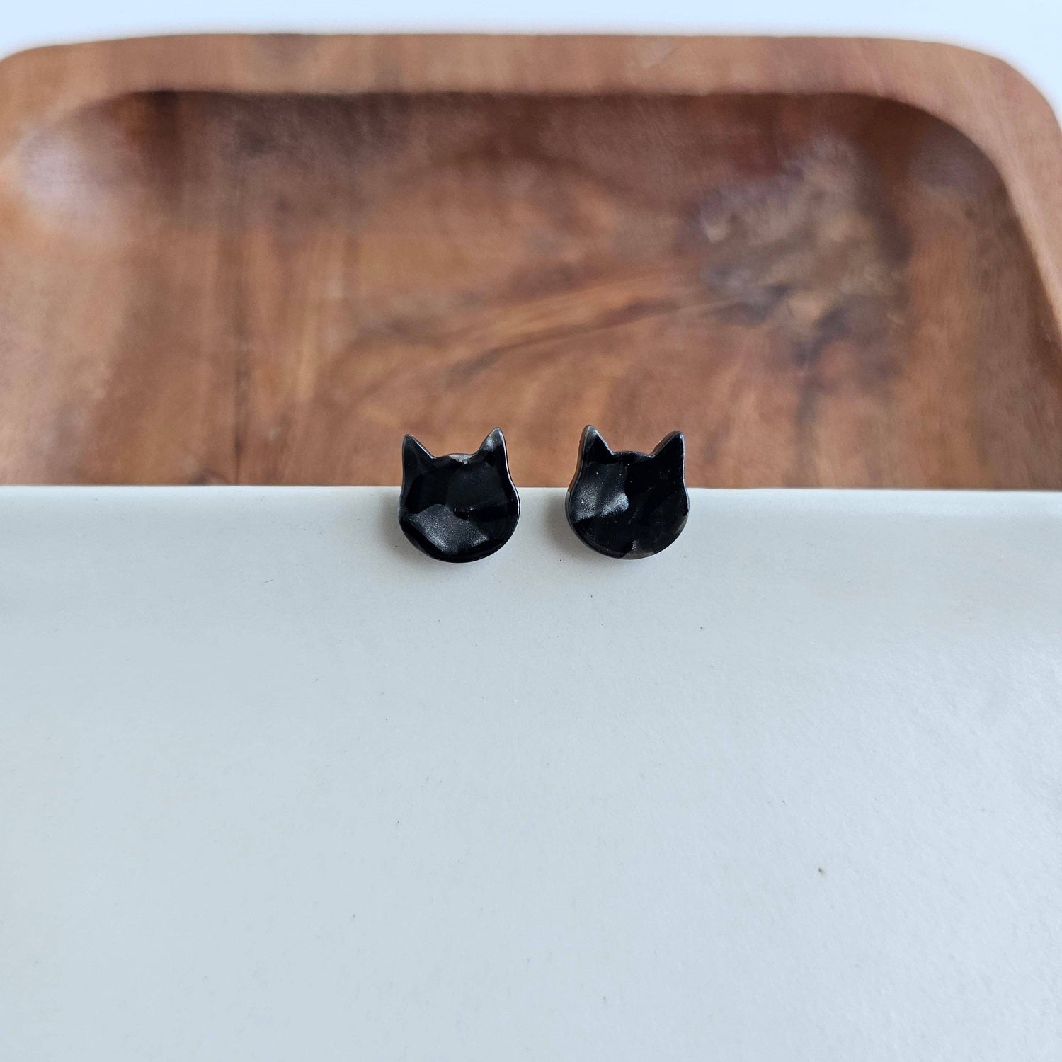 A pair of shiny black cat head stud earrings resting on a crisp white surface with a warm natural wooden bowl background