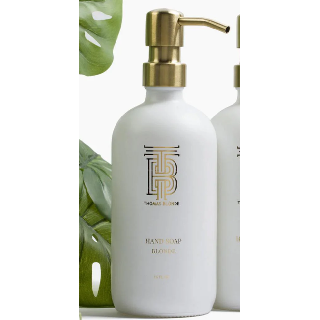 Product photography Thomas Blonde elegant white hand soap bottle with a brushed gold pump and gold monogram logo against a backdrop of green tropical monstera leaves