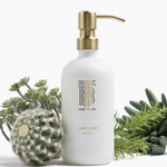 Thomas Blonde white hand lotion bottle with a gold pump and TB monogram logo sitting among various green succulents and a furry cactus