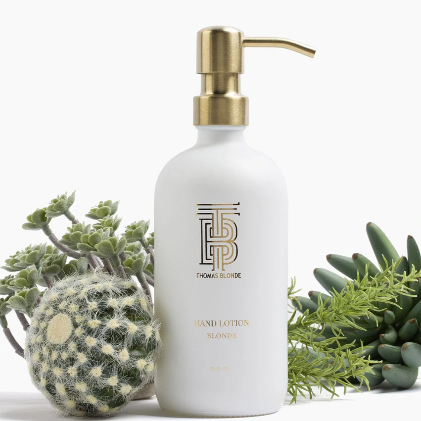 Thomas Blonde white hand lotion bottle with a gold pump and TB monogram logo sitting among various green succulents and a furry cactus