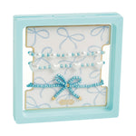 A Mud Pie set of three beaded bracelets displayed in a light blue frame, featuring white pearls, white heart beads, and a delicate blue beaded bow with gold accents on a bow-patterned background