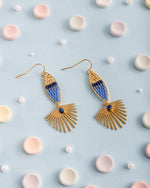 Handcrafted gold and ombre blue beaded drop earrings with geometric fan-shaped bottoms, arranged on a light blue surface among scattered pastel candies and small white pearls