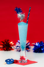 Blue frozen cocktail in a tall glass with a star patterned straw and metallic decorations on a red background