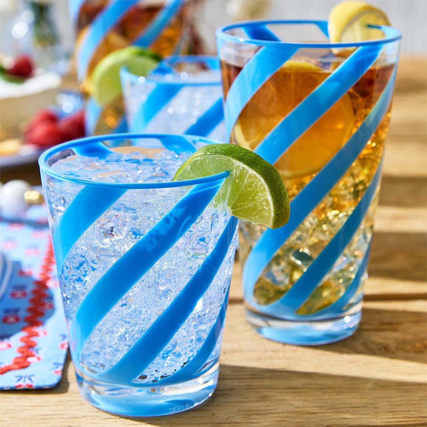 A photograph of blue-striped glassware filled with iced beverages on a sunny wooden table featuring swirling cobalt lines, carbonated bubbles in a clear drink, and fresh lime and lemon garnishes