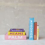 Colorful decorative books featuring titles like pink Palm, yellow Miami, purple Majorca, blue Mykonos, orange Matisse with a coral graphic, and cream Amalfi