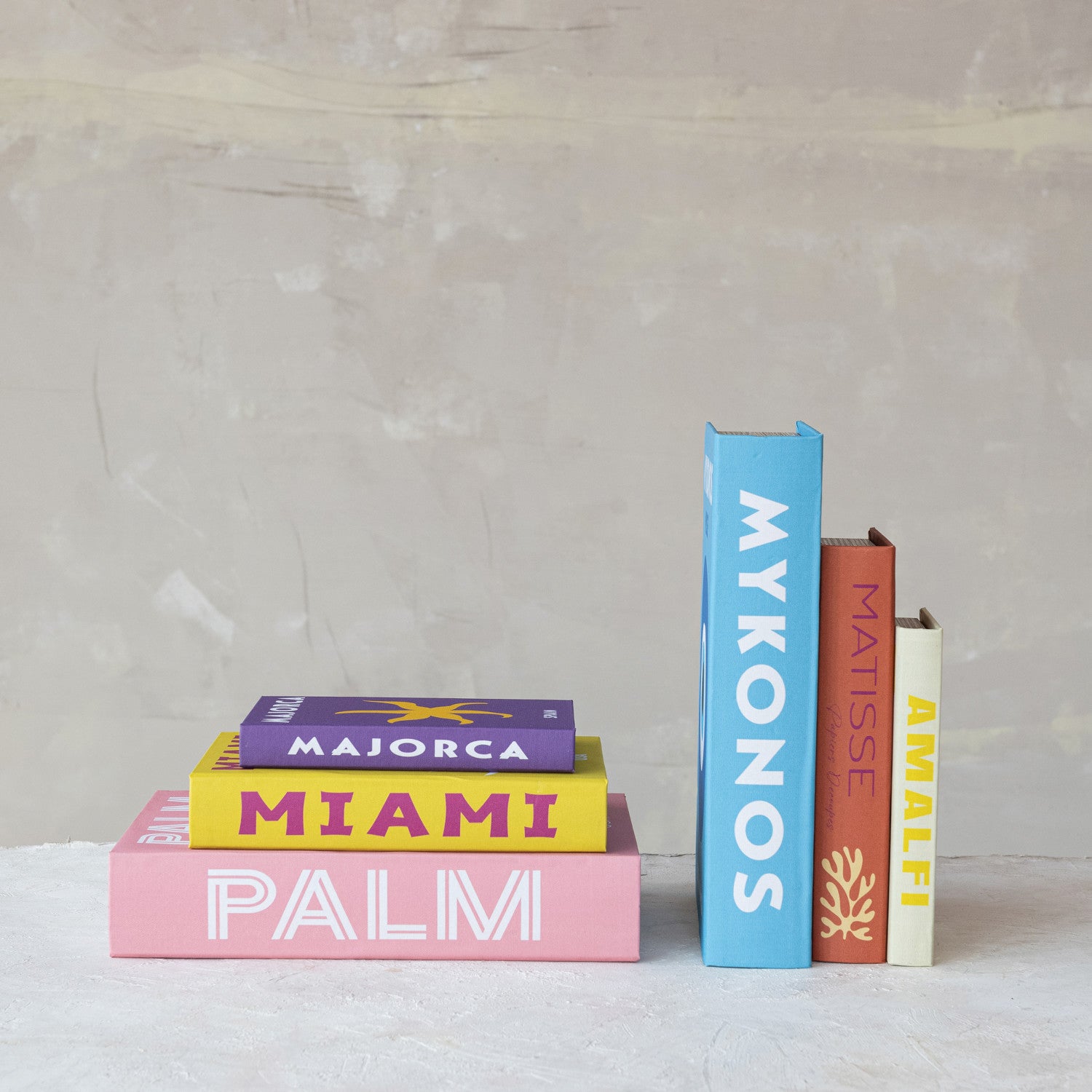 Colorful decorative books featuring titles like pink Palm, yellow Miami, purple Majorca, blue Mykonos, orange Matisse with a coral graphic, and cream Amalfi
