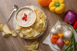 Bowl of creamy dip with red pepper garnish surrounded by potato chips and fresh vegetables on a wooden table