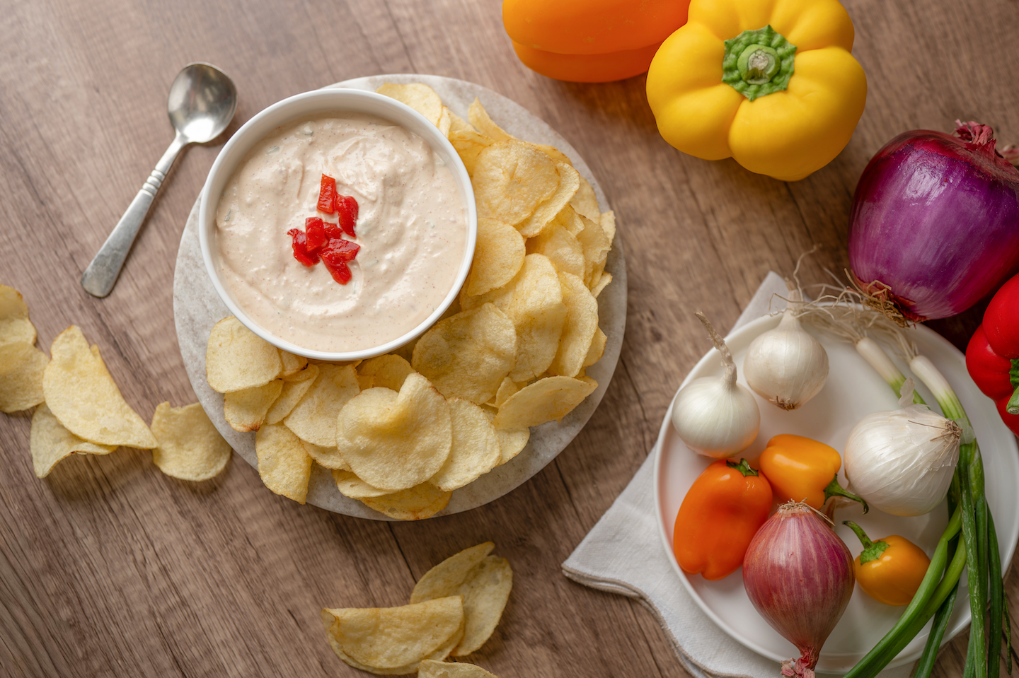 Bowl of creamy dip with red pepper garnish surrounded by potato chips and fresh vegetables on a wooden table