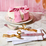 A celebratory white frosted cake adorned with a pink fondant bow and colorful sprinkles sits on a marble counter with gold bow-handled serving utensils on white embroidered napkins and two pink champagne flutes against a pink tiled wall
