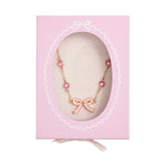 A delicate gold-toned necklace featuring a light pink enamel bow charm and five round pink gemstones presented in a light pink gift box with a scalloped window frame, white polka dot interior, and a white ribbon pull tab