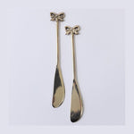 A pair of decorative gold-toned butter knives featuring intricate bow-shaped handles and slender polished blades