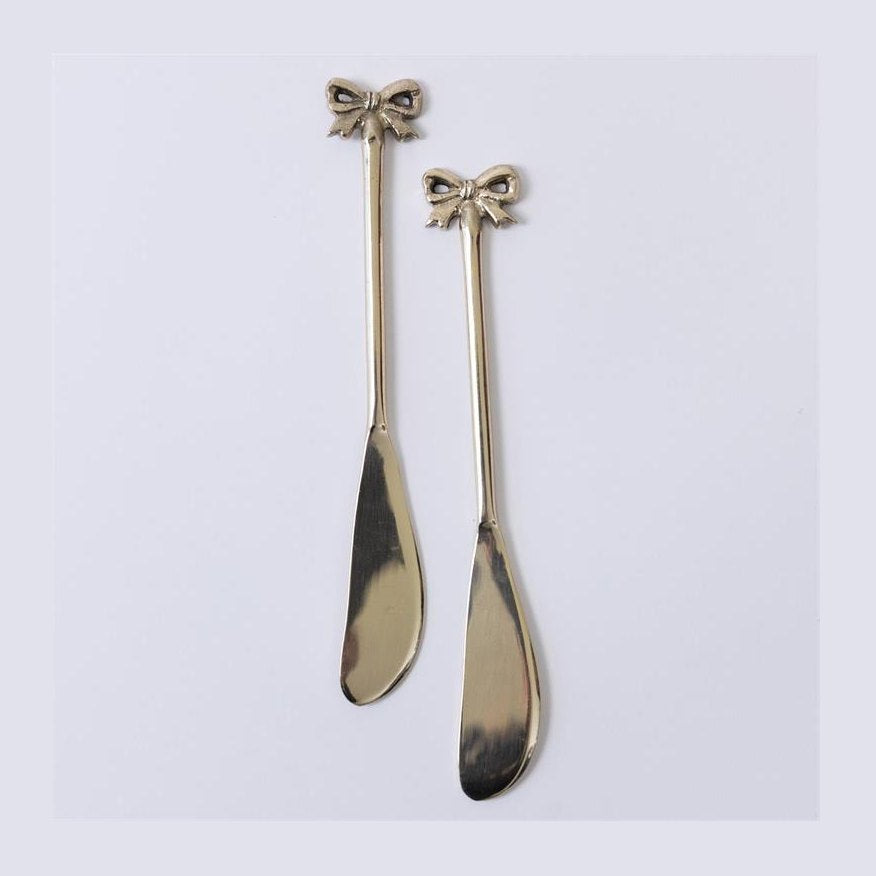 A pair of decorative gold-toned butter knives featuring intricate bow-shaped handles and slender polished blades