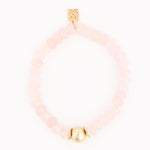 A pink beaded bracelet featuring smooth rose quartz beads a brushed gold spherical accent bead and a rectangular gold charm with an ornate geometric pattern on a clean white background