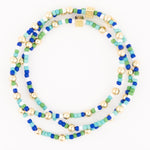 Beaded bracelets with small seed beads in royal blue, turquoise, green, and white, accented by polished gold spheres and two ornate etched square gold beads, coiled on a white background