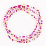 Three strands of fuchsia, pink, lavender, and white seed beads interspersed with gold spherical beads and square charms featuring an embossed cross-like pattern, arranged in an open circle