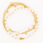 A coiled stack of gold and white beaded bracelets showcasing a square gold branded clasp a delicate gold cross charm and lustrous freshwater pearls
