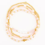 Photograph of layered beaded bracelets featuring gold seed beads, smooth blush beads, faceted pale pink crystals, and white pearls, accented with a small gold cross charm