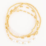 Three layered gold and white beaded bracelets featuring iridescent oval pearls, a small gold cross charm, and a cube bead with an etched symbol