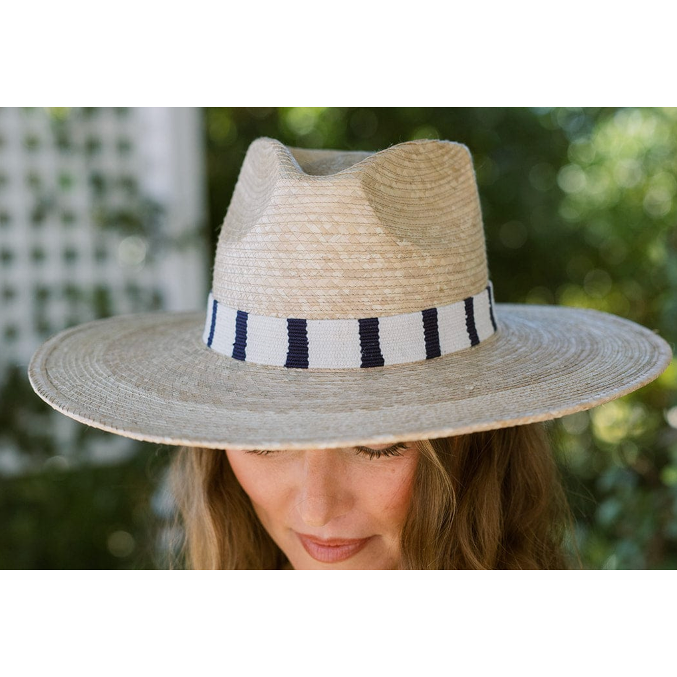 Photograph of a woman wearing a natural straw wide-brimmed hat with a white band featuring bold navy blue vertical stripes outdoors against soft green foliage and a white trellis