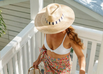 A woman in a wide-brimmed straw hat with a navy and white striped band, a white bikini top, and a vibrant floral sarong descends white wooden stairs carrying a woven straw bag