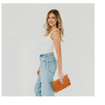 Photograph of a smiling blonde woman in a minimalist studio setting wearing a white tank top and light blue wide-leg jeans while carrying a textured cognac woven clutch bag