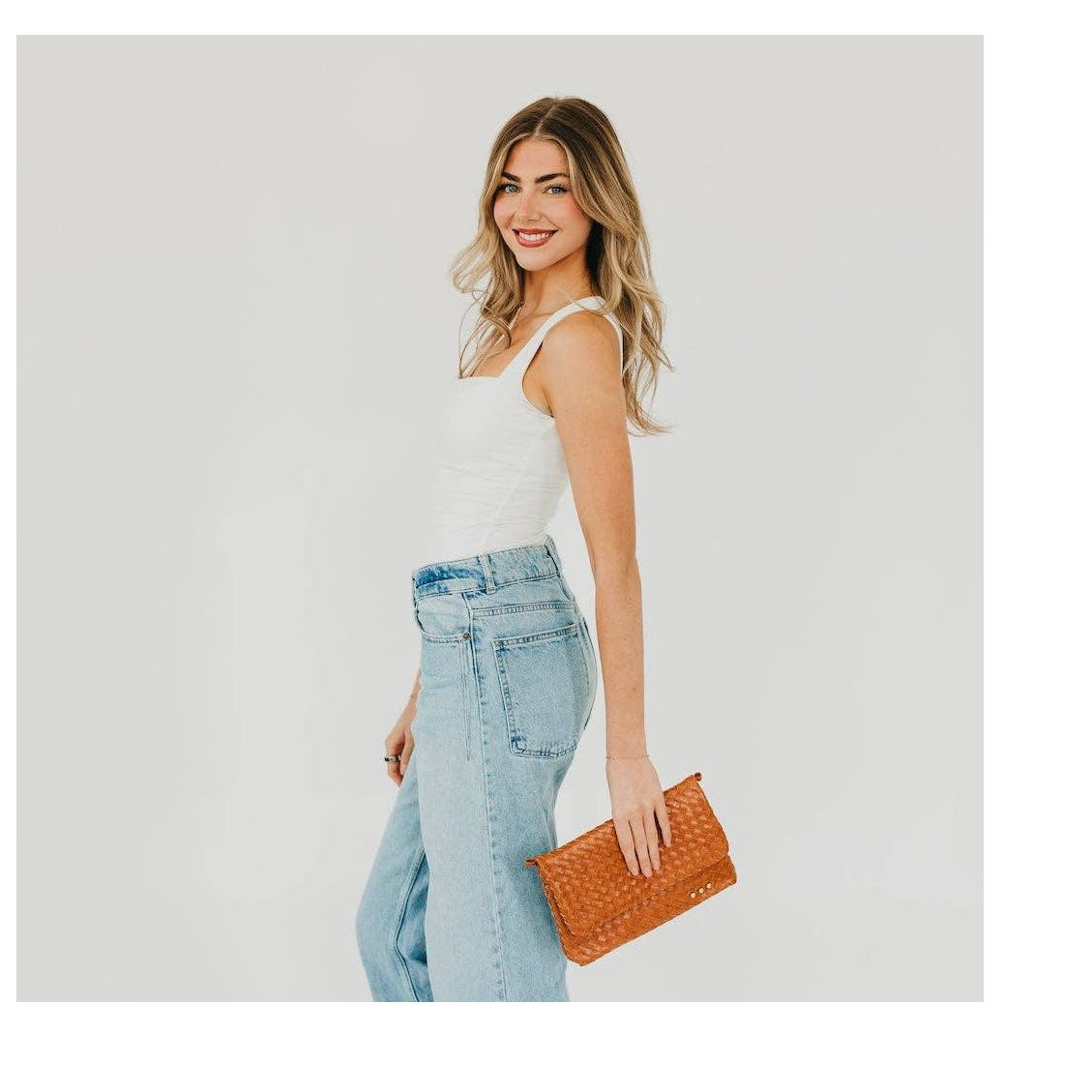 Photograph of a smiling blonde woman in a minimalist studio setting wearing a white tank top and light blue wide-leg jeans while carrying a textured cognac woven clutch bag