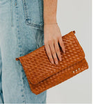 Photograph of a cognac-colored woven leather clutch held against light-wash denim jeans featuring an intricate basketweave texture, three small gold-tone studs, and a delicate gold chain bracelet