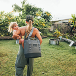 A woman in a camo cap carries a child with a heart band-aid on her knee and a gray YETI cooler bag through a lush tropical backyard