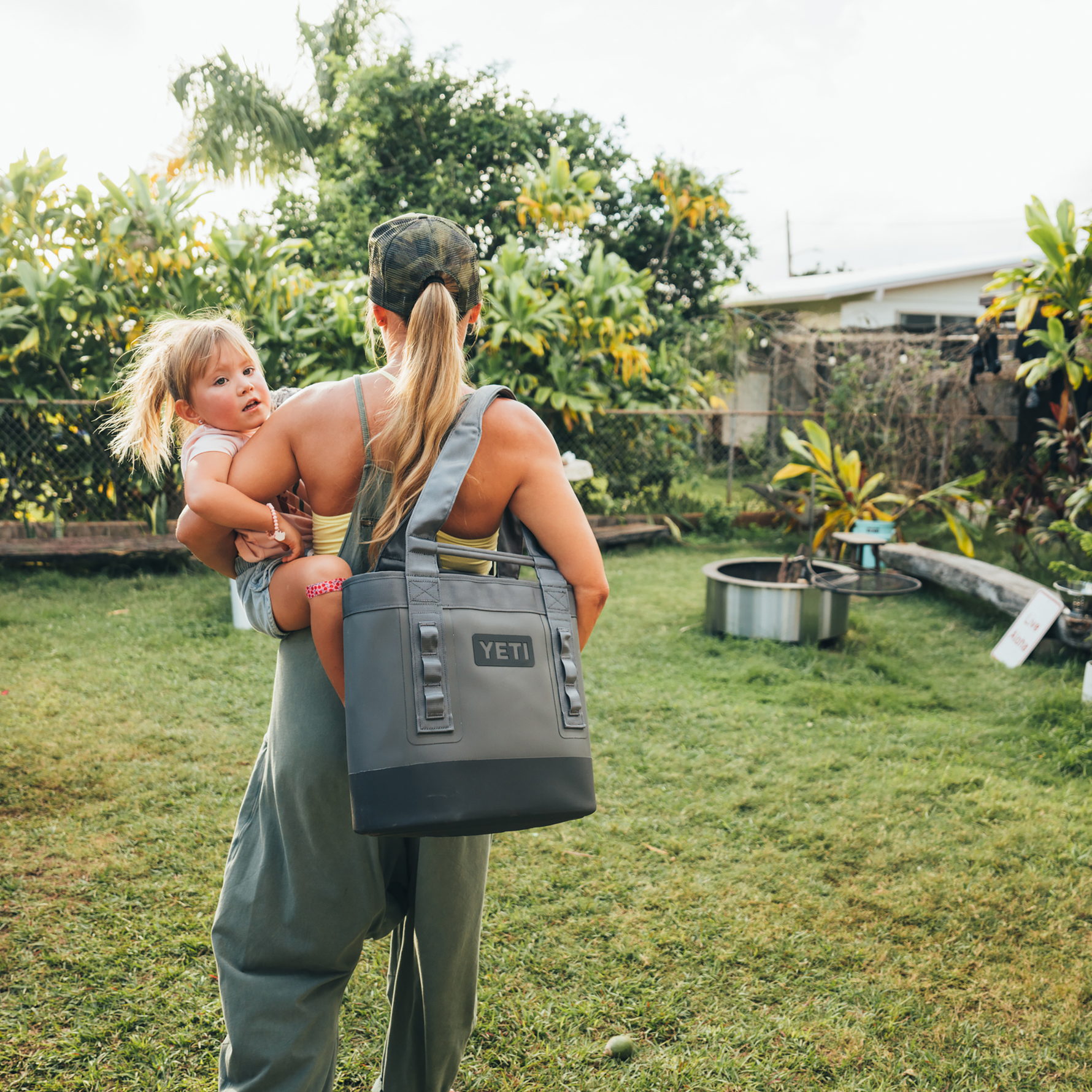 A woman in a camo cap carries a child with a heart band-aid on her knee and a gray YETI cooler bag through a lush tropical backyard