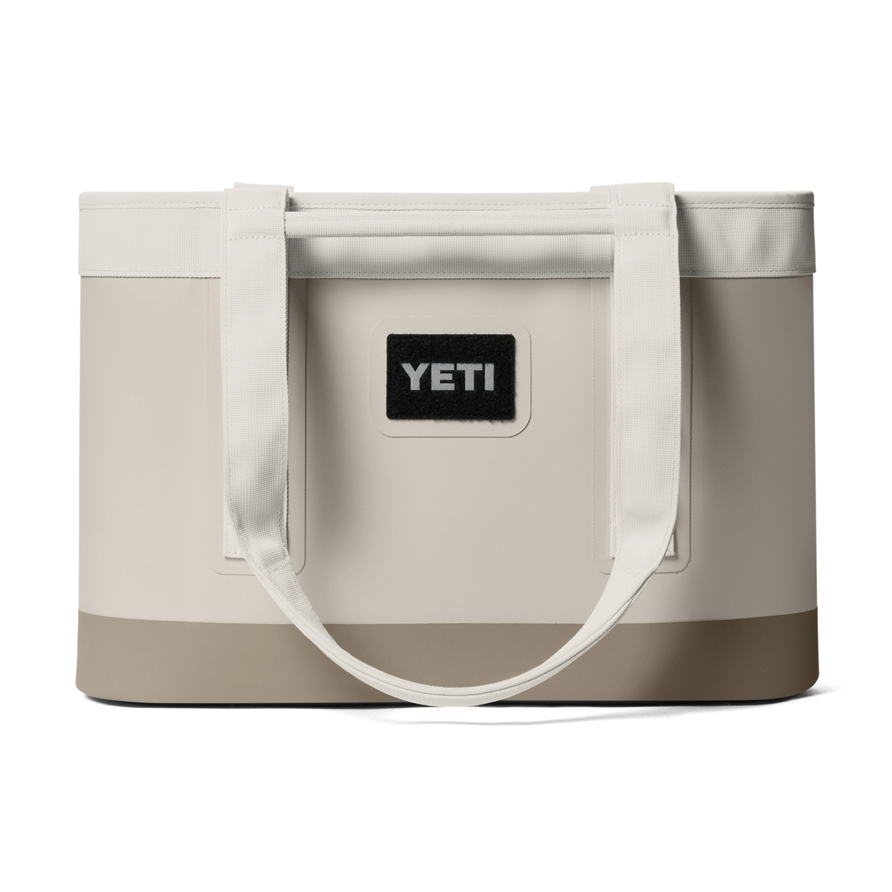 Product photography by YETI of a cream-colored Camino Carryall tote bag featuring a reinforced taupe base, thick woven shoulder straps, and a prominent black logo patch centered on the front against a dark background