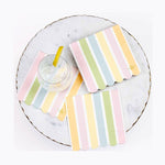 Pastel pink green blue and yellow striped napkins with scalloped edges and gold crown logos surround a clear drink with a lemon wedge on a white marble tray