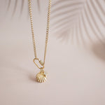 Photograph of a gold necklace featuring a ribbed seashell charm and a crystal star pendant attached to an oval carabiner clasp against a beige background with a soft palm leaf shadow