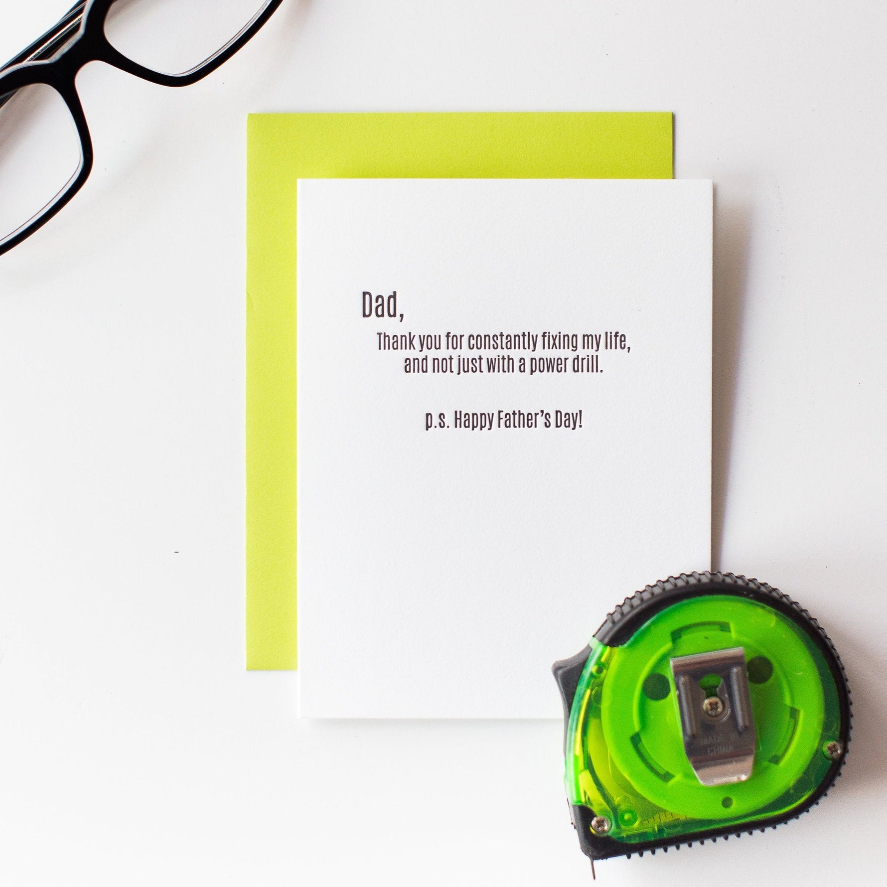 A flat lay of a Father's Day card with the message "Thank you for constantly fixing my life, and not just with a power drill" on a white surface alongside a bright lime green envelope, a fluorescent green and black tape measure, and black-rimmed eyeglasses