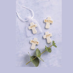 Photograph of four white ceramic crosses with gold lettering reading LOVE BLESSED JOY and FAITH arranged on a light blue textured surface with one cross in a sheer white drawstring bag and a green eucalyptus sprig