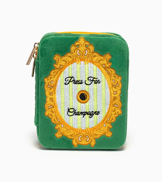 A vibrant green plush pouch with an elaborate gold embroidered oval frame containing black "Press For Champagne" text and a central black and gold button detail on a pale yellow and white striped background and a gold zipper