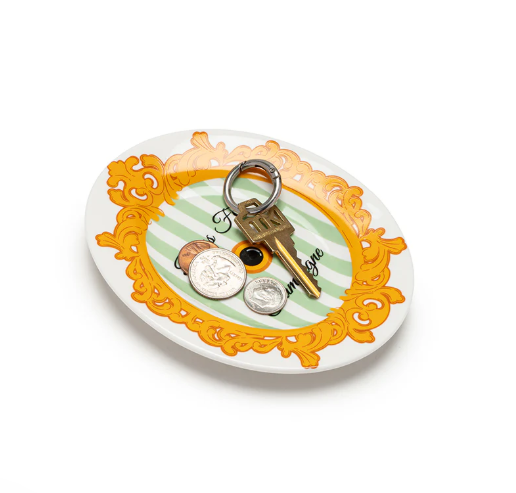 Oval ceramic trinket dish featuring a gold baroque border, green and white stripes, and partial "bonjour mon ami" text, holding a brass key, US quarter, dime, and penny on a white background