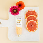 Dew Me hand cream tube with grapefruit slices and gerbera daisies on a marble board
