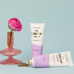 Two tubes of The Dream Cream hand creme with a pink flower in a glass vase and dried lavender on a blue background