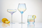 Product photography featuring three iridescent ribbed cocktail glasses including a coupe, a tall wine glass, and a tumblr with ice and a lemon twist, alongside a whole lemon and a wedge on a reflective white surface